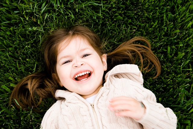 Smiling young girl with brown hair lying on green grass wearing a beige knitted sweater.