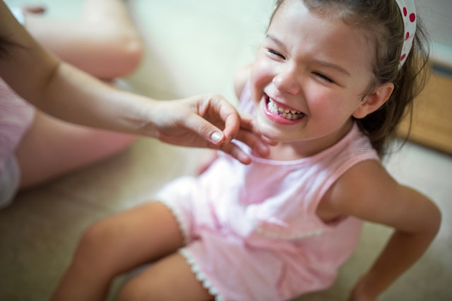 Smiling little girl wearing a pink dress, sitting on the floor, with a hand gently touching her chin.