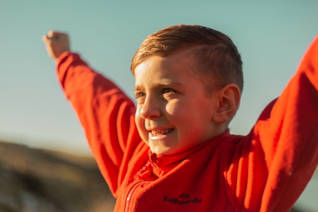 Smiling young boy in a red jacket raising his arms joyfully outdoors in sunlight.