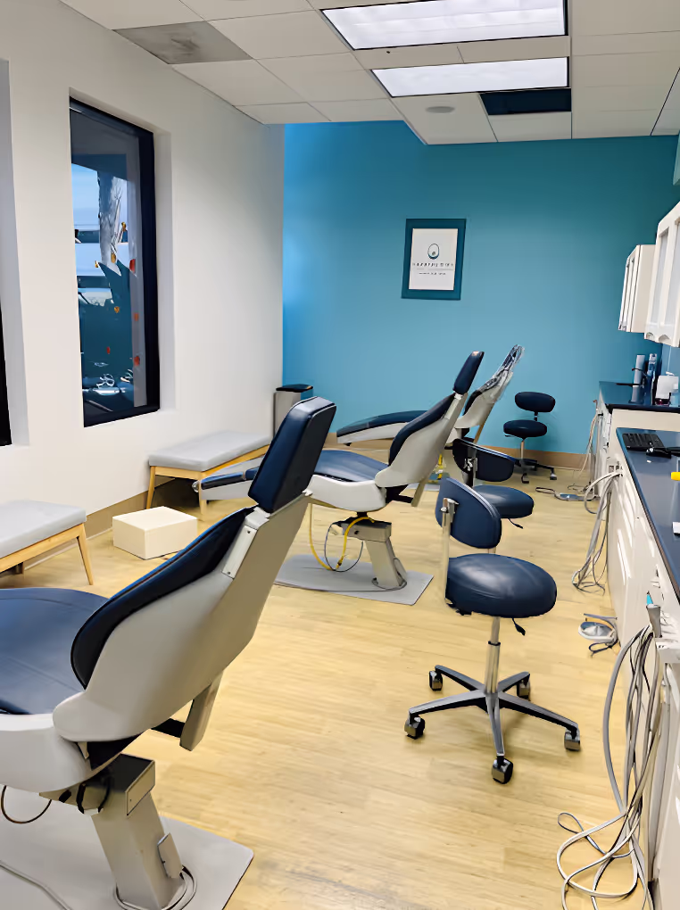 Modern dental office with three dental chairs, stools, blue accent wall, and wooden floor.