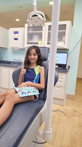 Smiling young girl sitting in a dental chair giving a thumbs up in a modern dental office.