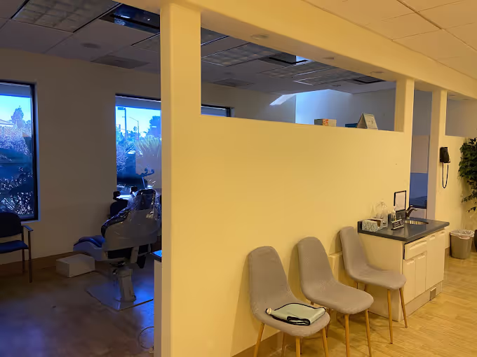 Interior of a dental office with three beige chairs, a sink countertop, and a dental chair visible in the background near windows.