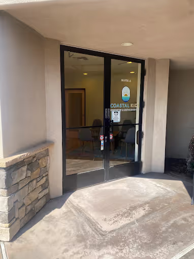 Glass double doors entrance to Coastal Kids Pediatric Dentistry and Orthodontics with stone wall on the left and concrete walkway.