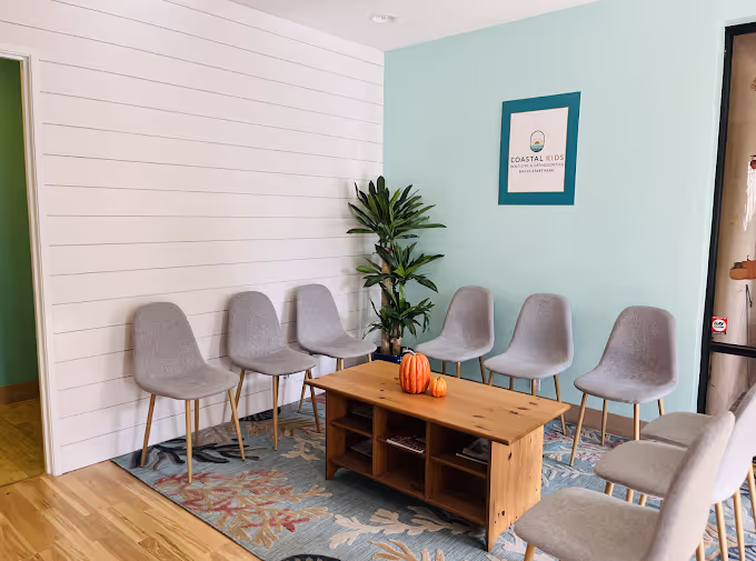 Waiting room with gray chairs arranged in a semicircle, a wooden coffee table with two small pumpkins, a green plant in the corner, and a Coastal Kids sign on a light blue wall.