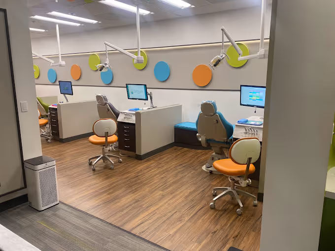 Modern dental clinic with patient chairs, orange stools, computer screens, and colorful wall-mounted circles.