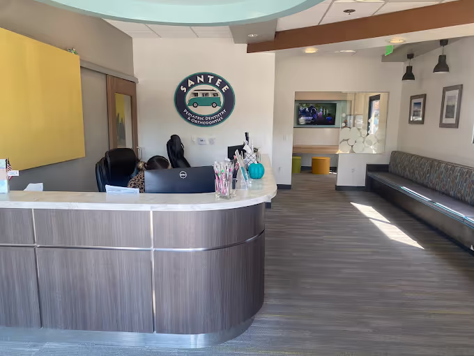 Reception area of Santee Pediatric Dentistry and Orthodontics with a curved wooden desk, two receptionists, seating along the right wall, and a logo sign on the back wall.