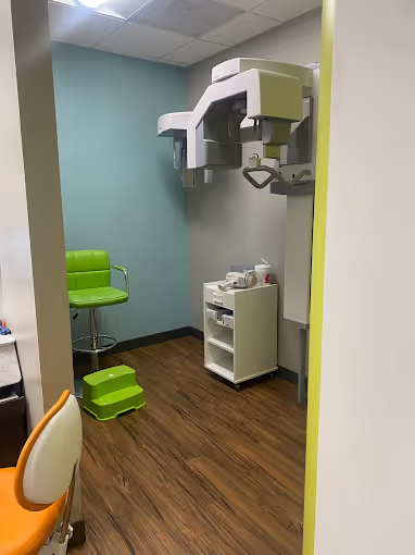 Dental X-ray room with green chair, green footstool, dental imaging machine, and a small white cart with supplies.