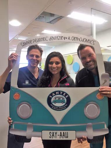 Three people smiling behind a photo frame shaped like a teal and white van with text 'Santee Pediatric Dentistry & Orthodontics' and license plate 'Say-Aah'.
