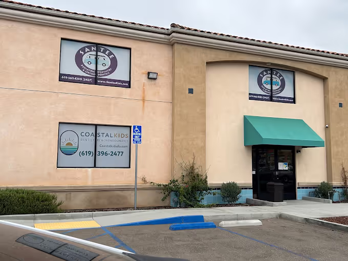 Exterior view of a beige commercial building with signs for Santee Pediatric Dentistry and Coastal Kids orthodontics, featuring a green awning above the entrance and handicapped parking spaces in front.