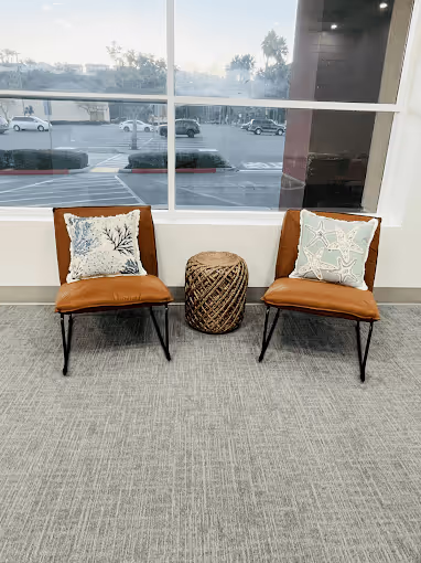 Two brown leather chairs with patterned cushions and a woven basket between them in front of a large window overlooking a parking lot.