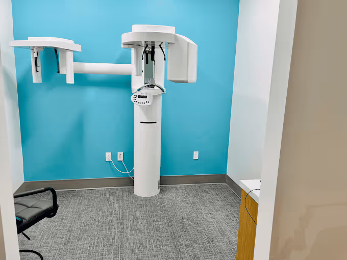 A dental panoramic X-ray machine in a small examination room with a blue accent wall, gray carpet, and a black office chair.