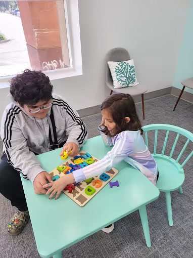 An adult and a child sitting at a small turquoise table playing with colorful wooden alphabet letters on a board.