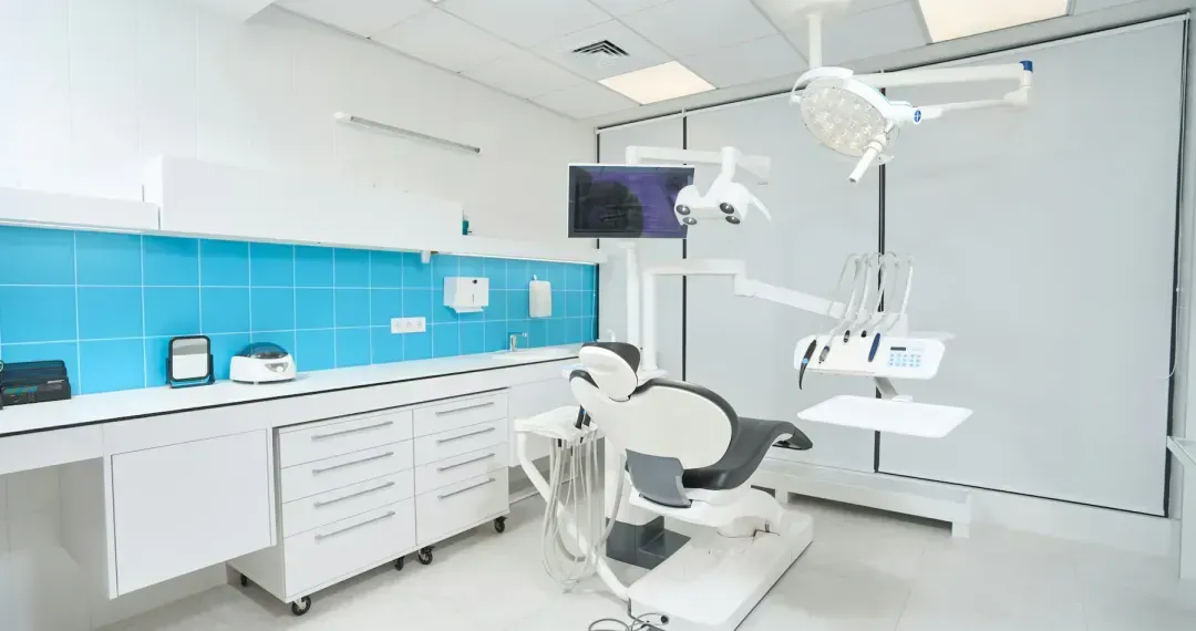 Bright, modern dental clinic room with a dental chair, overhead light, equipment, and blue tiled backsplash.