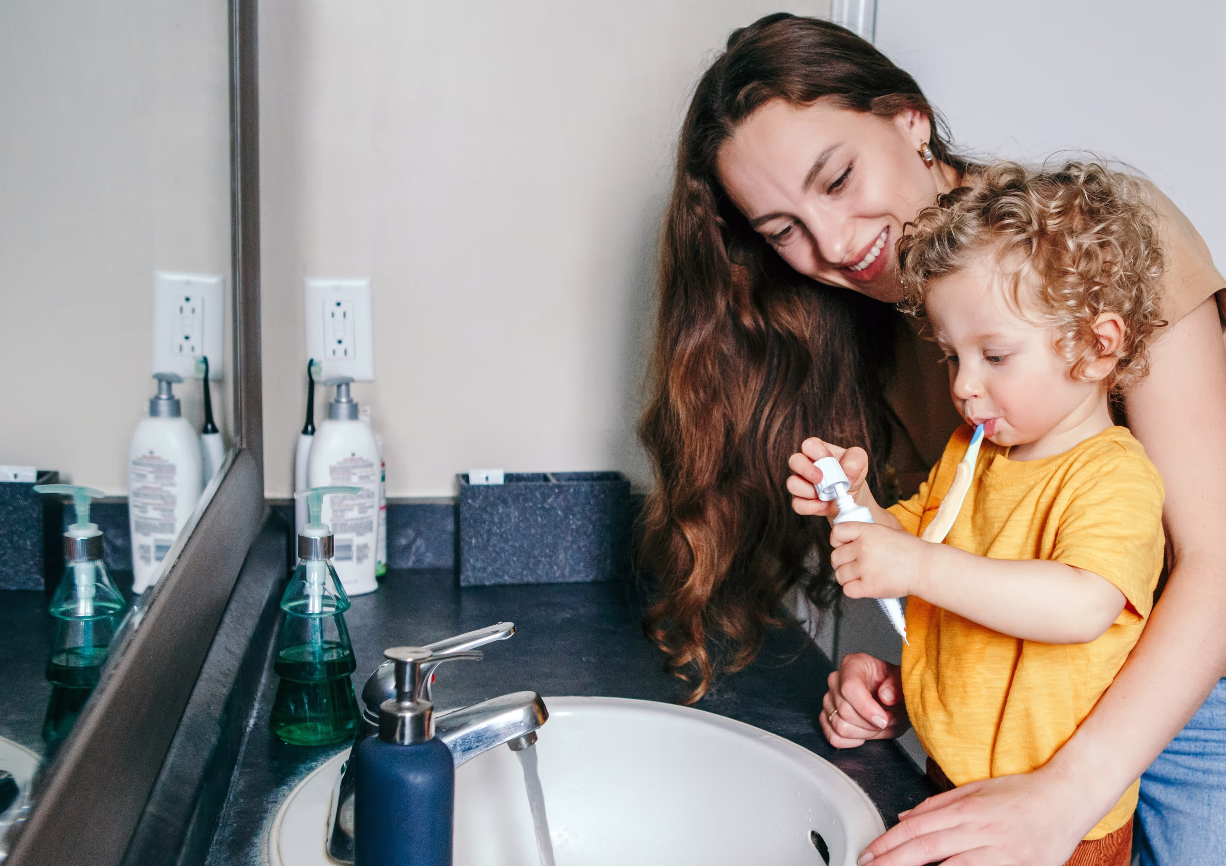 Mother helping young child put toothpaste on toothbrush at bathroom sink with running water.
