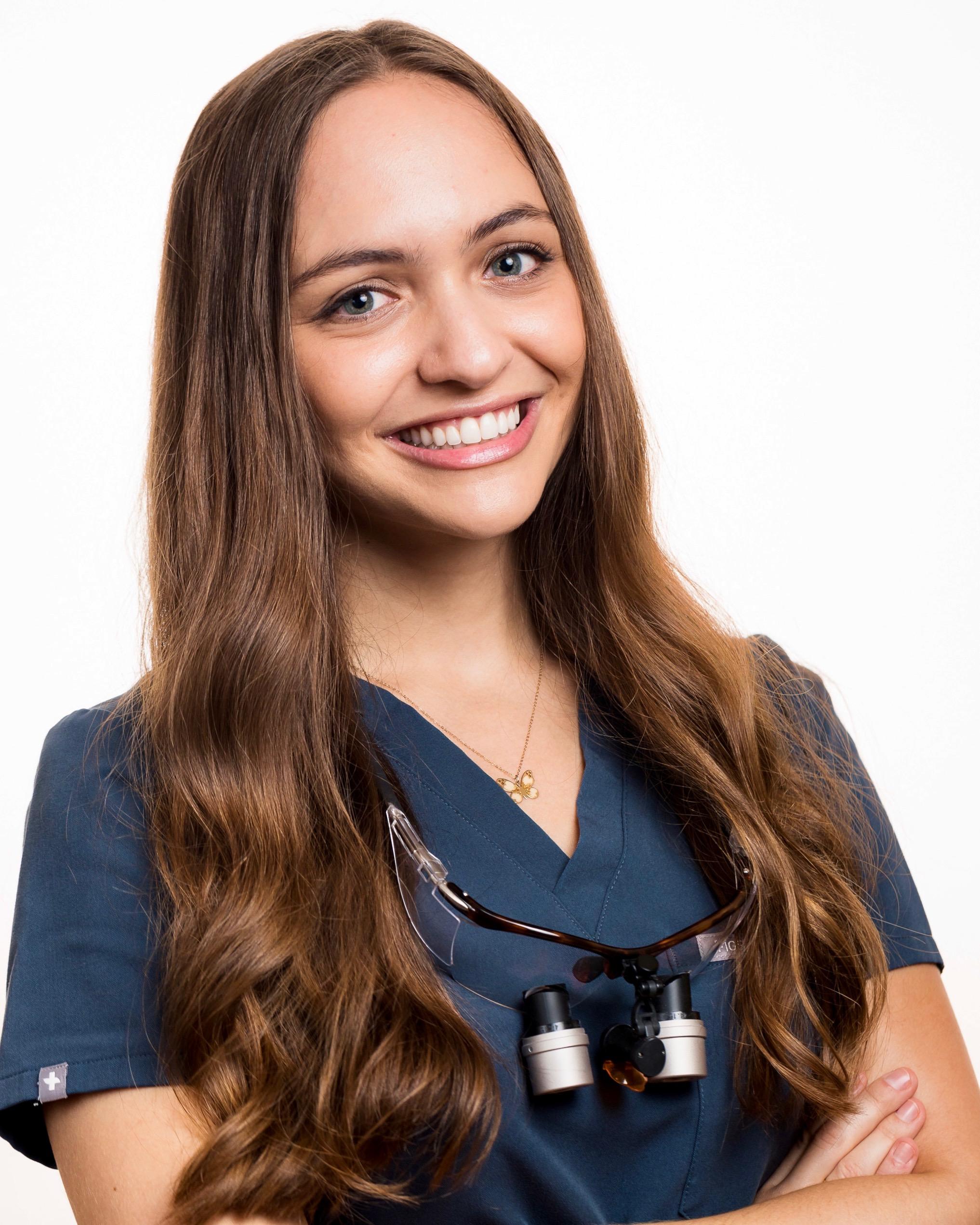 Smiling woman with long brown hair wearing navy scrubs and dental loupes, arms crossed.