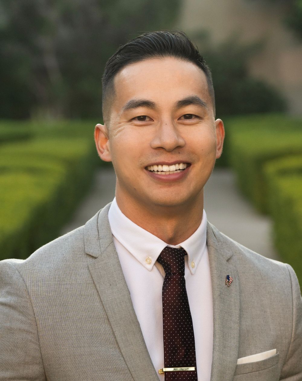 Smiling man in a light gray suit with a white shirt and dark polka dot tie, outdoors with greenery background.