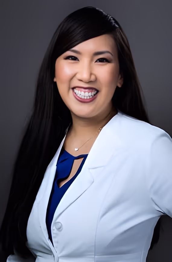 Smiling woman with long black hair wearing a white lab coat and blue blouse against a dark background.